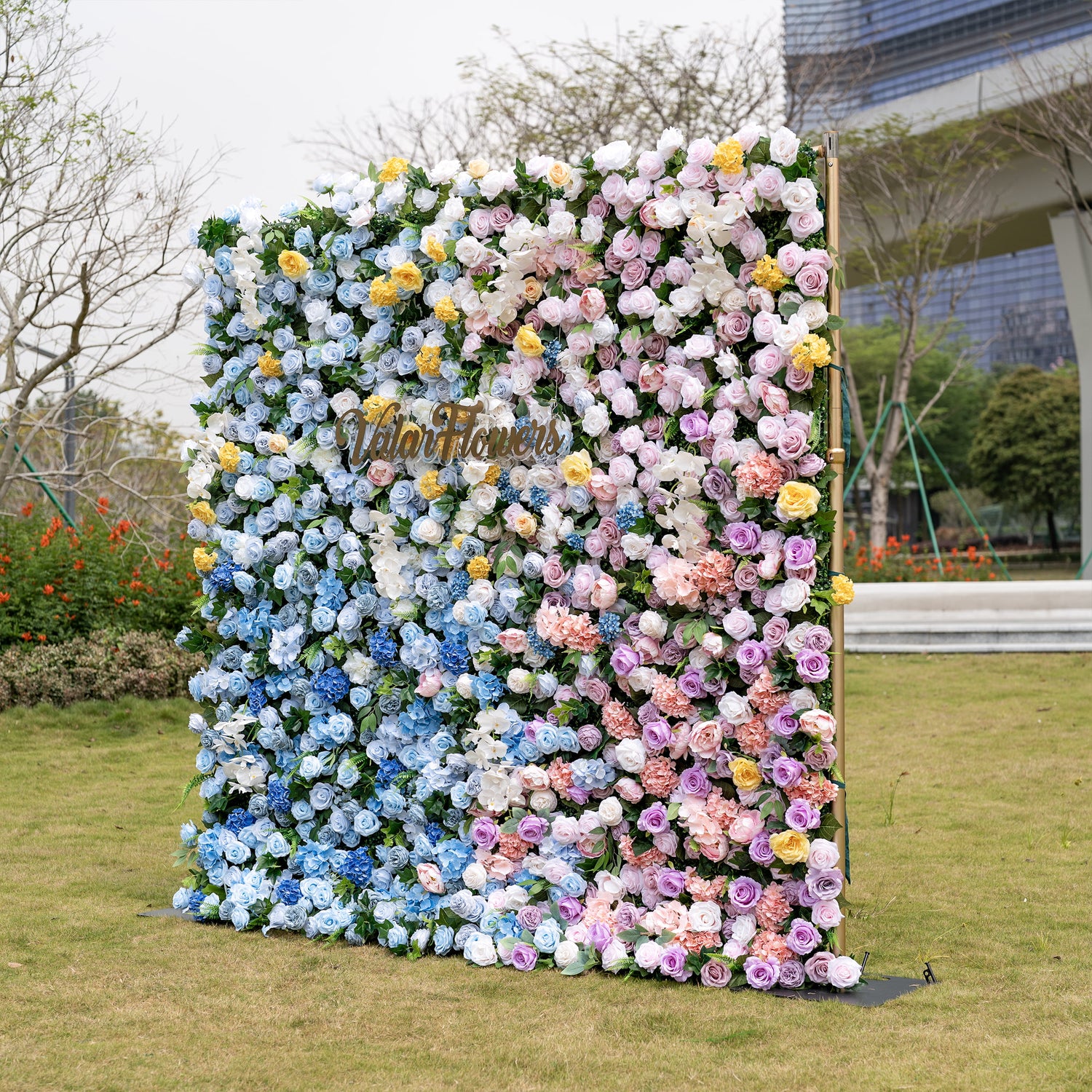 This is a stunning gradient artificial flower wall backdrop from Valar Flowers, set outdoors on a grassy lawn with a modern urban skyline in the background. It features a lush, dense arrangement of lifelike blooms: soft sky-blue delphiniums at the bottom, transitioning upward to creamy white roses, sunny yellow poppies, blush pink peonies, and lavender roses. The seamless color gradient creates an elegant, romantic focal point perfect for weddings, photo booths, and luxury event decor.