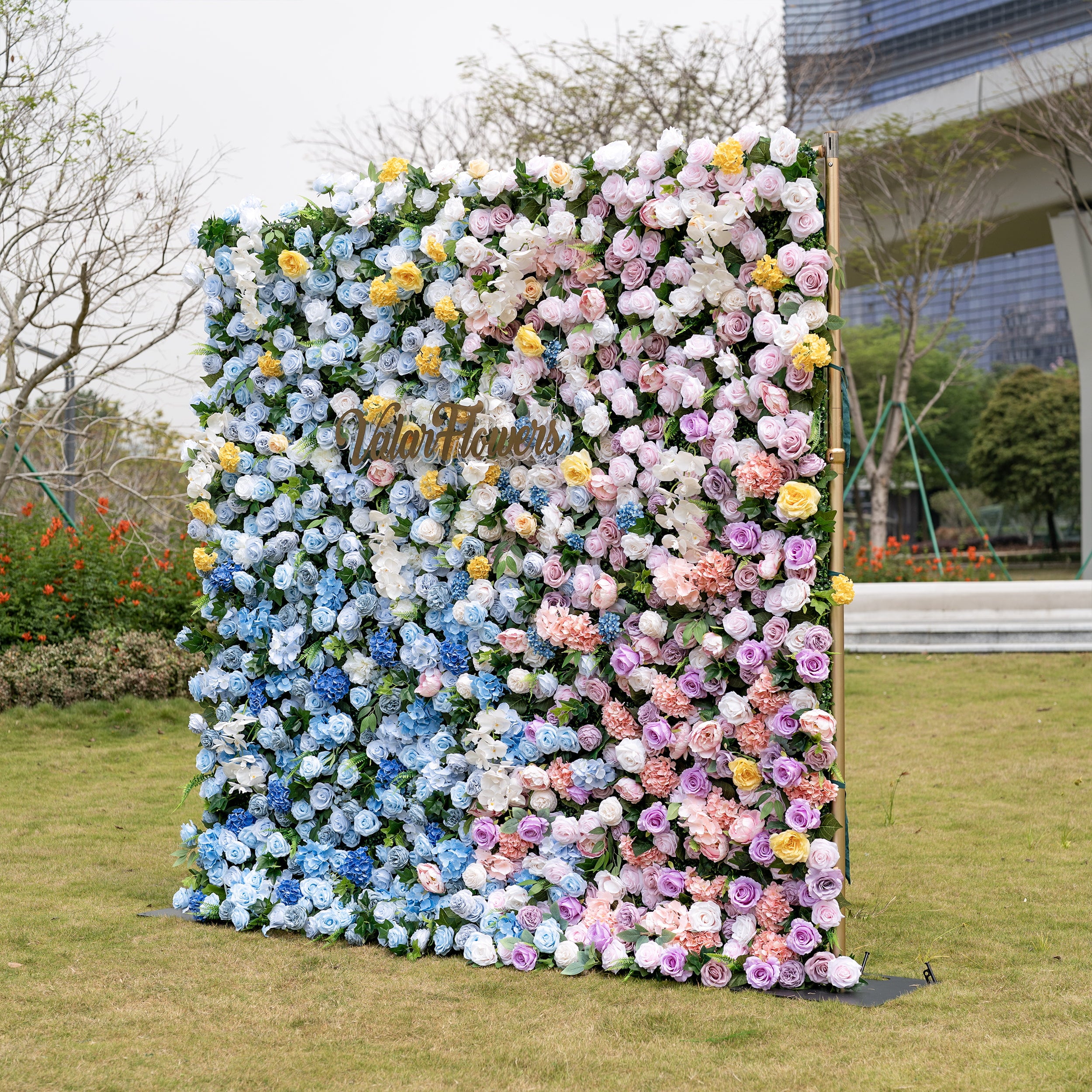 This is a stunning gradient artificial flower wall backdrop from Valar Flowers, set outdoors on a grassy lawn with a modern urban skyline in the background. It features a lush, dense arrangement of lifelike blooms: soft sky-blue delphiniums at the bottom, transitioning upward to creamy white roses, sunny yellow poppies, blush pink peonies, and lavender roses. The seamless color gradient creates an elegant, romantic focal point perfect for weddings, photo booths, and luxury event decor.