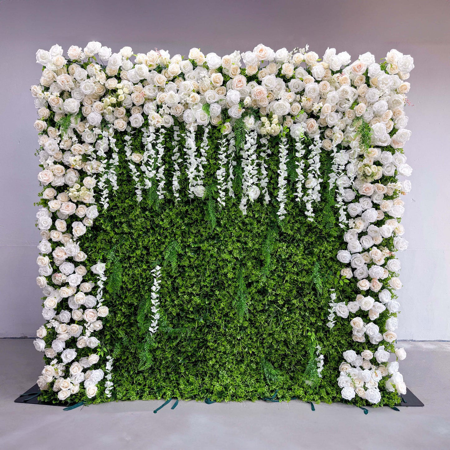 This flower wall has a green leafy background with a profusion of white roses. The roses are clustered together, especially at the top and sides. Hanging white floral strands add a dynamic element, contributing to the wall's sophisticated and romantic appearance.