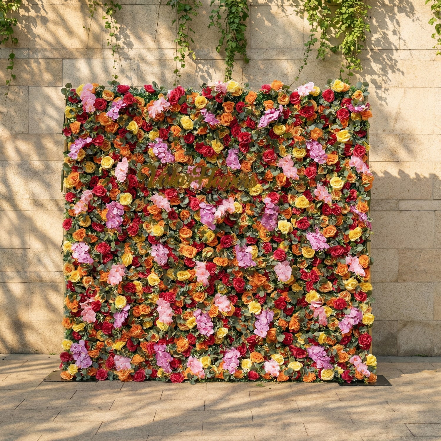 This is a vibrant, full-size artificial flower wall backdrop from ValarFlowers, set against a sunlit stone wall with trailing ivy. It features a lush, dense arrangement of lifelike blooms in a bright, cheerful multicolor palette: red, orange, yellow, pink, and lavender roses, plus delicate pink filler flowers and green foliage. This premium, reusable floral display creates a bold, romantic focal point, perfect for summer weddings, garden parties, photo booths, and luxury outdoor event decor.