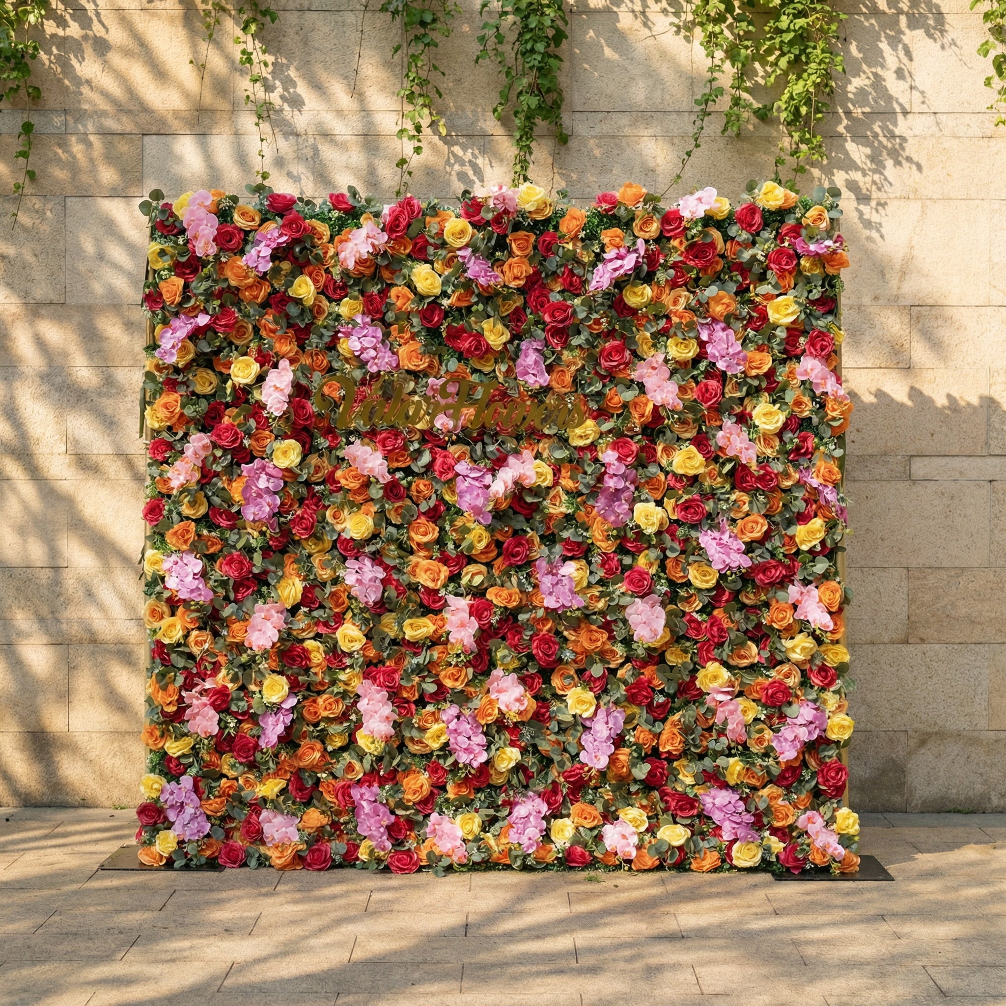 This is a vibrant, full-size artificial flower wall backdrop from ValarFlowers, set against a sunlit stone wall with trailing ivy. It features a lush, dense arrangement of lifelike blooms in a bright, cheerful multicolor palette: red, orange, yellow, pink, and lavender roses, plus delicate pink filler flowers and green foliage. This premium, reusable floral display creates a bold, romantic focal point, perfect for summer weddings, garden parties, photo booths, and luxury outdoor event decor.