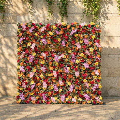 This is a vibrant, full-size artificial flower wall backdrop from ValarFlowers, set against a sunlit stone wall with trailing ivy. It features a lush, dense arrangement of lifelike blooms in a bright, cheerful multicolor palette: red, orange, yellow, pink, and lavender roses, plus delicate pink filler flowers and green foliage. This premium, reusable floral display creates a bold, romantic focal point, perfect for summer weddings, garden parties, photo booths, and luxury outdoor event decor.