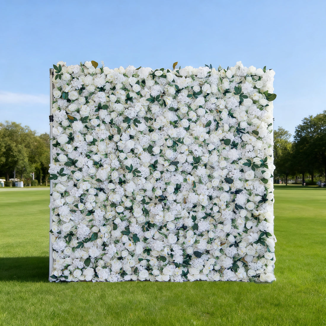 This image features a large, square floral wall densely covered with white flowers, interspersed with green leaves. Set outdoors on a lush green lawn under a clear blue sky, with trees in the background, it creates an elegant and serene atmosphere. Though &quot;Valar Flowers&quot; branding isn&