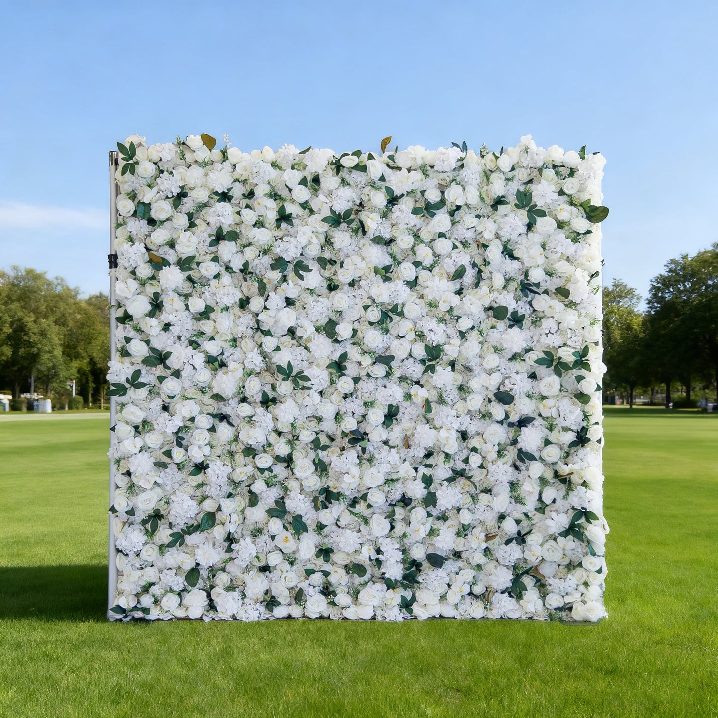 This image features a large, square floral wall densely covered with white flowers, interspersed with green leaves. Set outdoors on a lush green lawn under a clear blue sky, with trees in the background, it creates an elegant and serene atmosphere. Though "Valar Flowers" branding isn't visible here, the display is perfect for events or decorative purposes, exuding a fresh and refined charm.