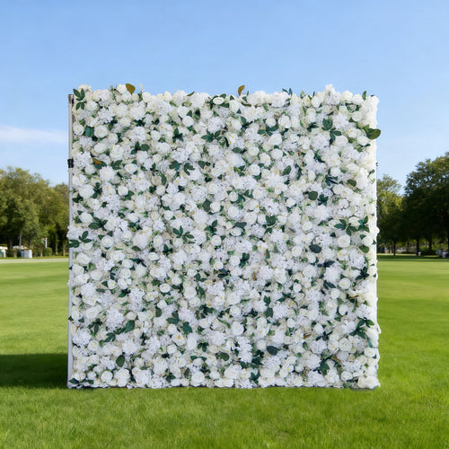 This image features a large, square floral wall densely covered with white flowers, interspersed with green leaves. Set outdoors on a lush green lawn under a clear blue sky, with trees in the background, it creates an elegant and serene atmosphere. Though "Valar Flowers" branding isn't visible here, the display is perfect for events or decorative purposes, exuding a fresh and refined charm.