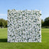 This image features a large, square floral wall densely covered with white flowers, interspersed with green leaves. Set outdoors on a lush green lawn under a clear blue sky, with trees in the background, it creates an elegant and serene atmosphere. Though "Valar Flowers" branding isn&