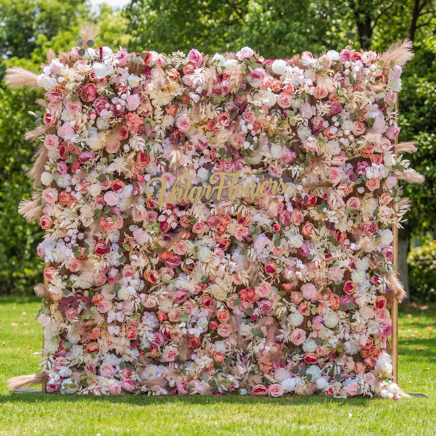 This outdoor floral backdrop (set on lush green grass, tree-framed) blends soft pinks, whites, and deep rose blooms with airy feathers, creating a romantic, textured display. A polished, cursive "Valar Flowers" gold sign anchors the center, contrasting with the lush, multi-tonal arrangement. The elegant, dreamy palette exudes warmth and sophistication, perfect for wedding decor, garden events, or upscale photo setups.