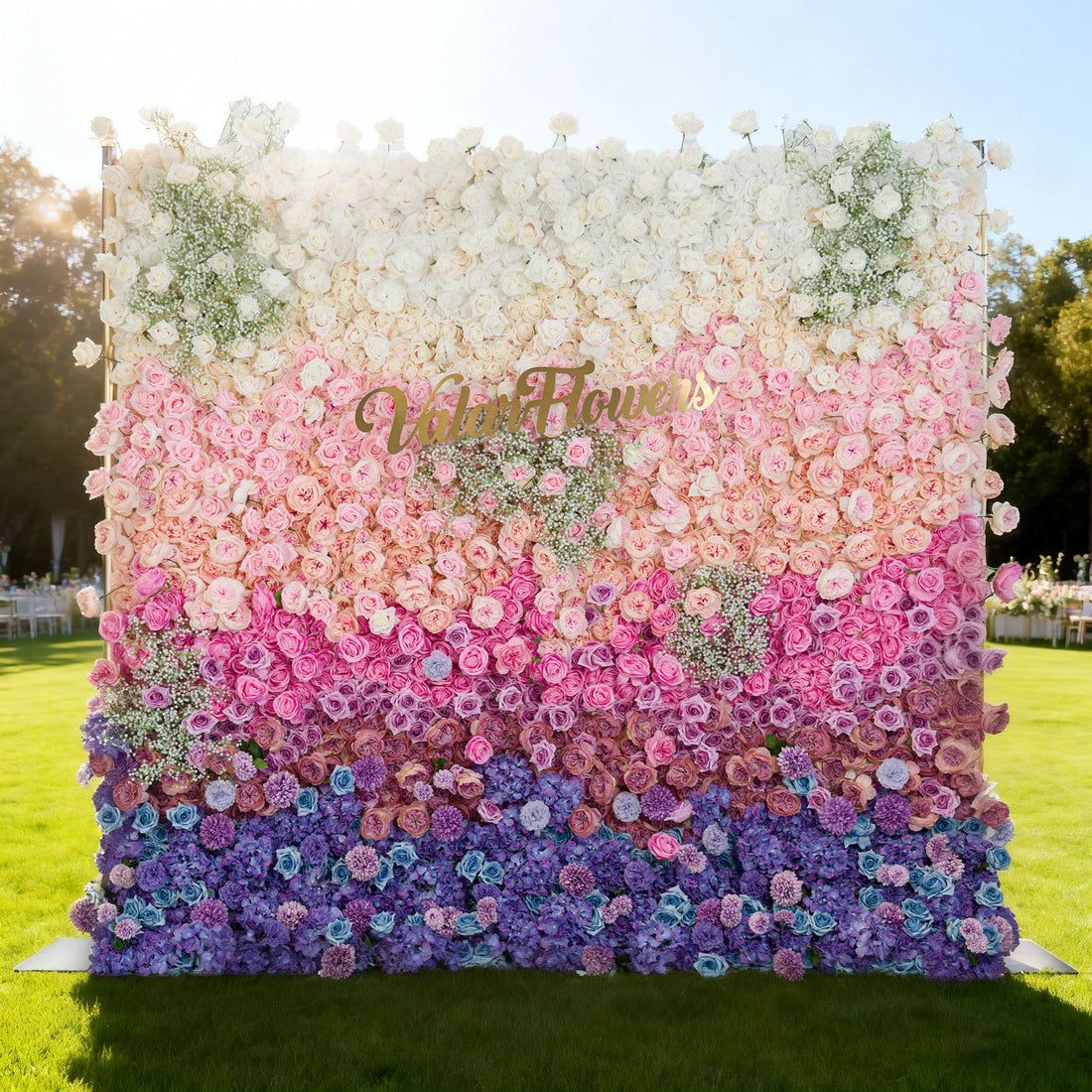 This image features a large floral wall with a stunning gradient of colors, transitioning from white and pink at the top to purple and blue at the bottom. &quot;Valar Flowers&quot; is displayed in golden text. Set outdoors on a lush green lawn under a bright sky, with event setups in the background, it creates a vibrant and elegant atmosphere, perfect for special occasions.