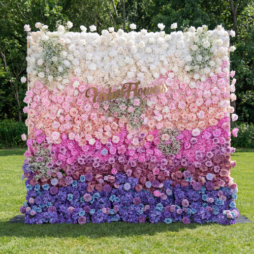 A stunning gradient floral wall by Valar Flowers stands on lush green grass, transitioning from creamy white at the top through soft pink, deep magenta, and rich purple, to vibrant blue at the bottom. Densely packed with roses, peonies, and baby's breath, it features a gold brand sign, creating a luxurious, eye-catching backdrop ideal for outdoor weddings, brand events, or photo shoots.