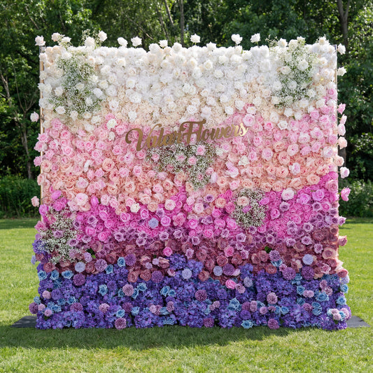 A stunning gradient floral wall by Valar Flowers stands on lush green grass, transitioning from creamy white at the top through soft pink, deep magenta, and rich purple, to vibrant blue at the bottom. Densely packed with roses, peonies, and baby's breath, it features a gold brand sign, creating a luxurious, eye-catching backdrop ideal for outdoor weddings, brand events, or photo shoots.