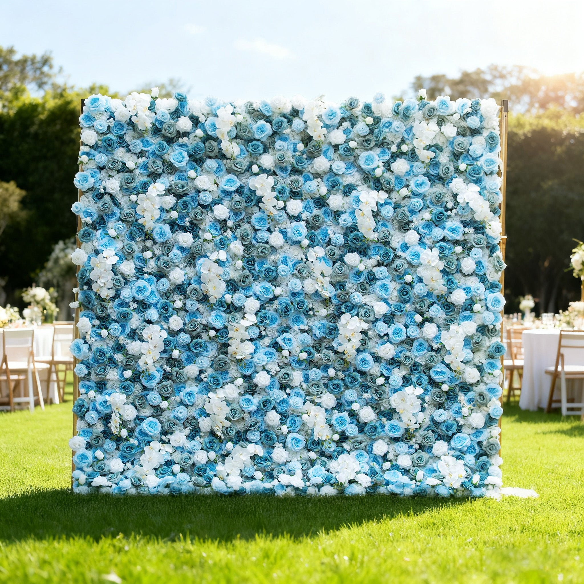 This image showcases a beautiful floral wall by Valar Flowers, set outdoors on a lush green lawn. It features a dense arrangement of artificial flowers in various shades of blue and white, creating a serene and elegant display. The backdrop is perfect for events, with the soft hues and lush texture adding a touch of sophistication, complemented by the sunny outdoor setting and event furniture in the background.