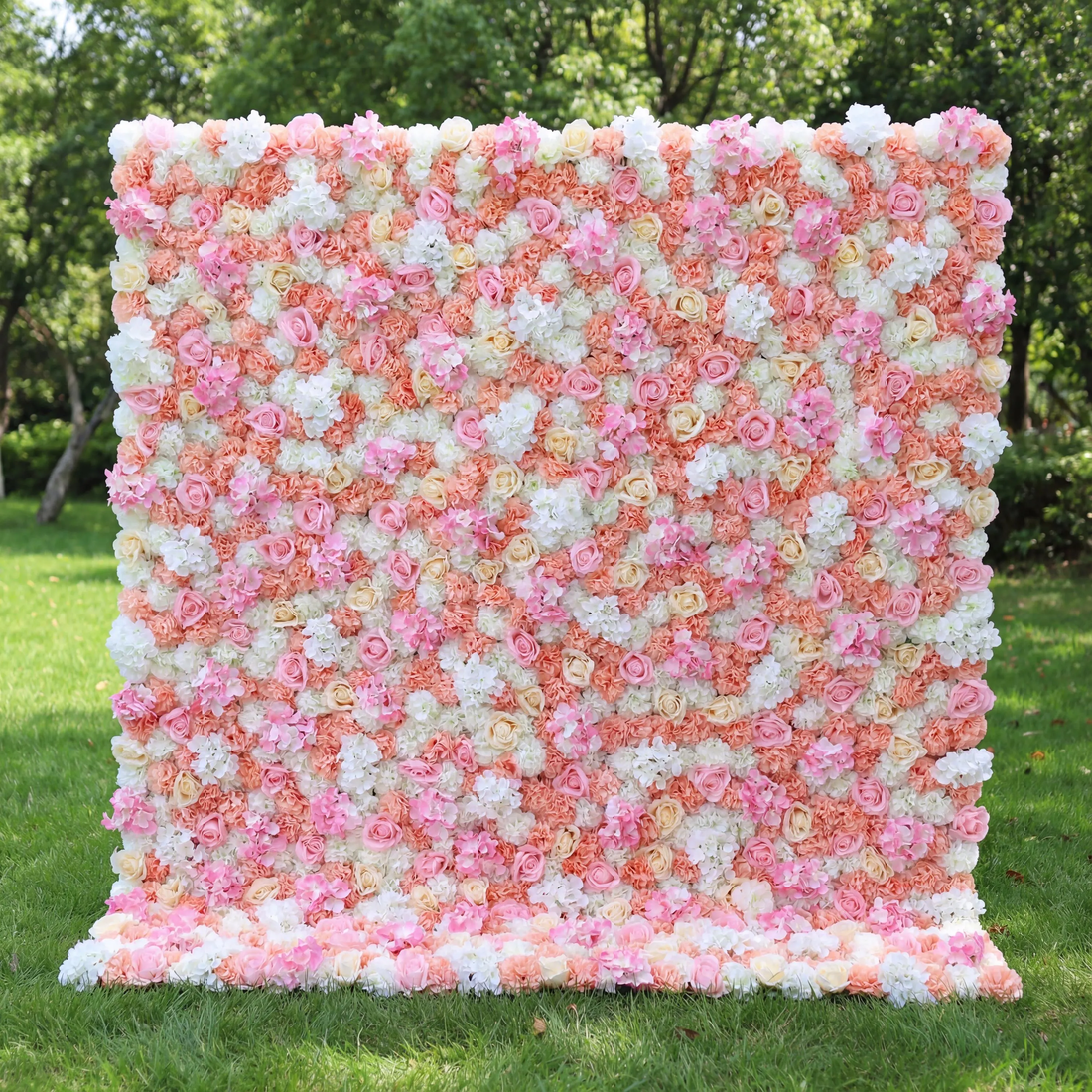 This vibrant floral wall (set on sunlit grass, tree backdrop) blends soft pinks, peachy corals, and crisp whites, layered in a dense, textured scatter of blooms. Natural light amplifies the warm, cheerful palette, enhancing the airy, playful vibe. Ideal for festive events or celebrations, it serves as an eye-catching backdrop—merging bright, inviting color with lush, romantic charm to feel both lively and elegant.