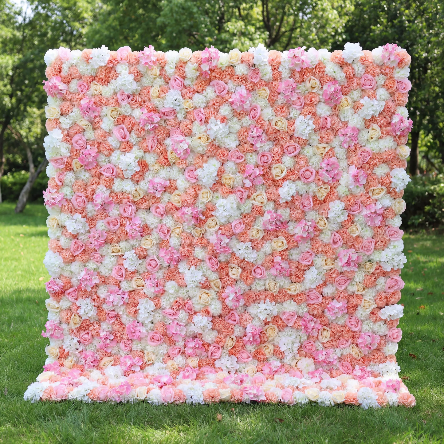 This vibrant floral wall (set on sunlit grass, tree backdrop) blends soft pinks, peachy corals, and crisp whites, layered in a dense, textured scatter of blooms. Natural light amplifies the warm, cheerful palette, enhancing the airy, playful vibe. Ideal for festive events or celebrations, it serves as an eye-catching backdrop—merging bright, inviting color with lush, romantic charm to feel both lively and elegant.