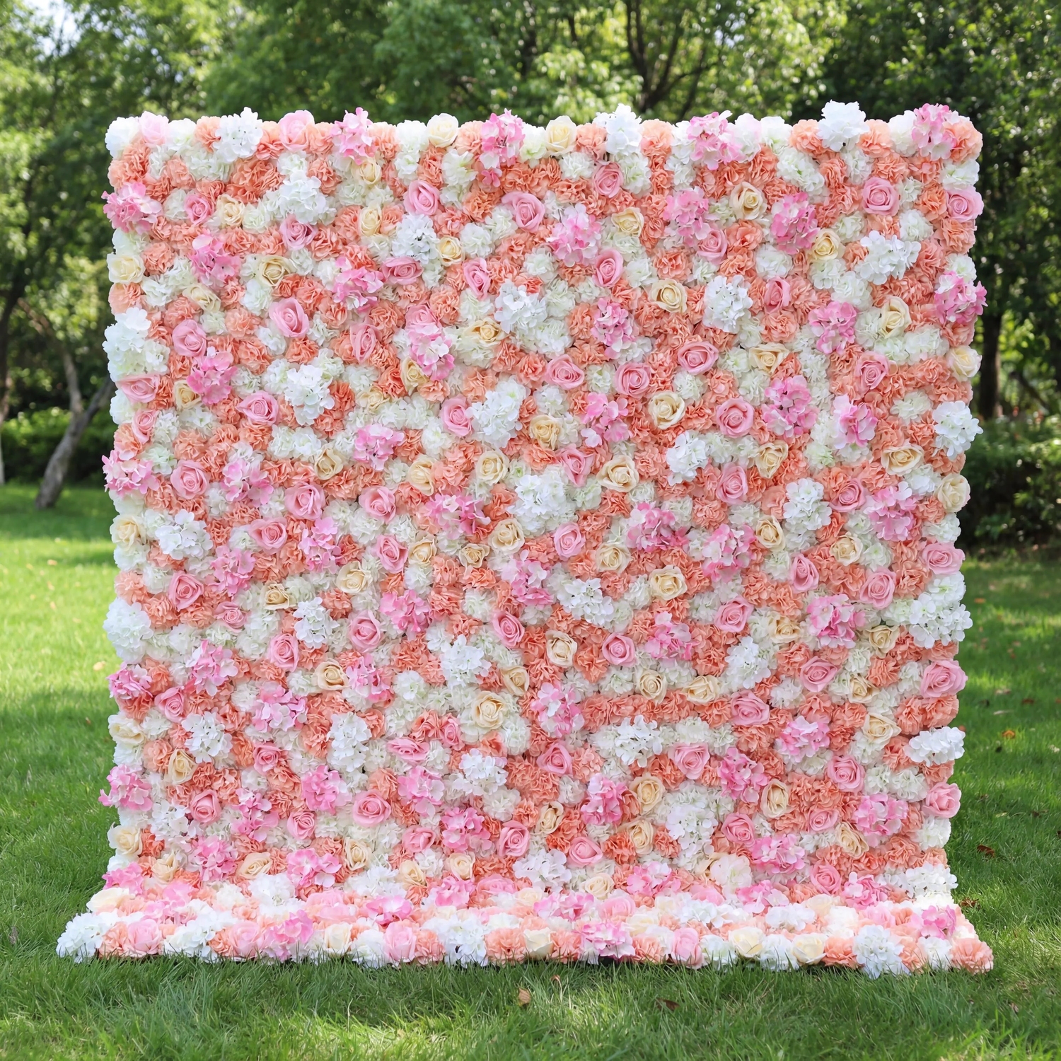 This vibrant floral wall (set on sunlit grass, tree backdrop) blends soft pinks, peachy corals, and crisp whites, layered in a dense, textured scatter of blooms. Natural light amplifies the warm, cheerful palette, enhancing the airy, playful vibe. Ideal for festive events or celebrations, it serves as an eye-catching backdrop—merging bright, inviting color with lush, romantic charm to feel both lively and elegant.