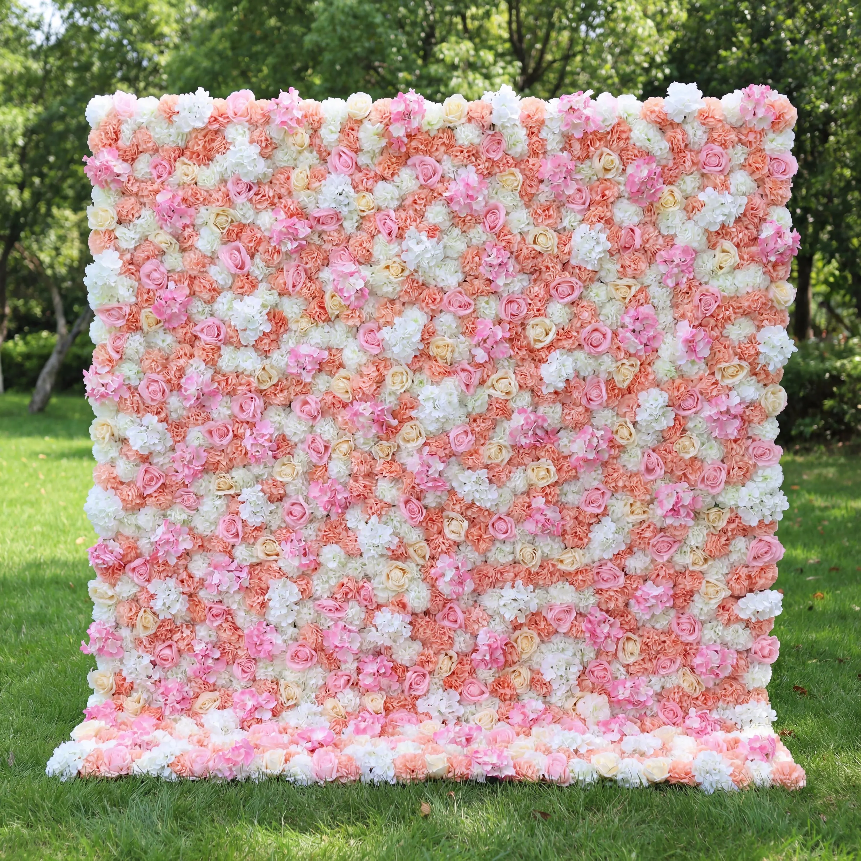This vibrant floral wall (set on sunlit grass, tree backdrop) blends soft pinks, peachy corals, and crisp whites, layered in a dense, textured scatter of blooms. Natural light amplifies the warm, cheerful palette, enhancing the airy, playful vibe. Ideal for festive events or celebrations, it serves as an eye-catching backdrop—merging bright, inviting color with lush, romantic charm to feel both lively and elegant.