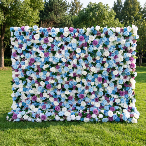 Dreamy blue artificial flower wall densely packed with sky-blue roses, creamy white blooms, vivid purple pom-pom flowers and subtle green foliage, set on a bright sunlit lawn with tall green trees. This reusable, low-upkeep floral backdrop delivers cool whimsical tones, ideal for baby boy showers, gender reveal parties, wedding photo booths and blue-themed social media shoots for refreshing, eye-catching celebration decor.