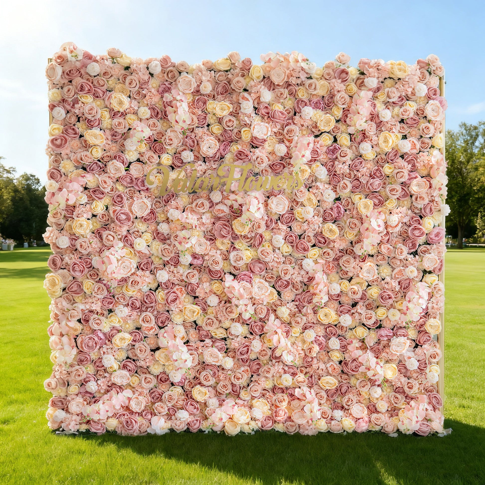 This image features a large, square floral wall outdoors on a lush green lawn. It's densely covered with an array of flowers in soft hues like pink, peach, and cream. The sign "Valar Flowers" is prominently displayed on the floral wall. Bathed in sunlight, with trees and a clear blue sky in the background, the scene exudes a sense of beauty, elegance, and natural charm, perfect for decorative purposes in events or as a picturesque backdrop.