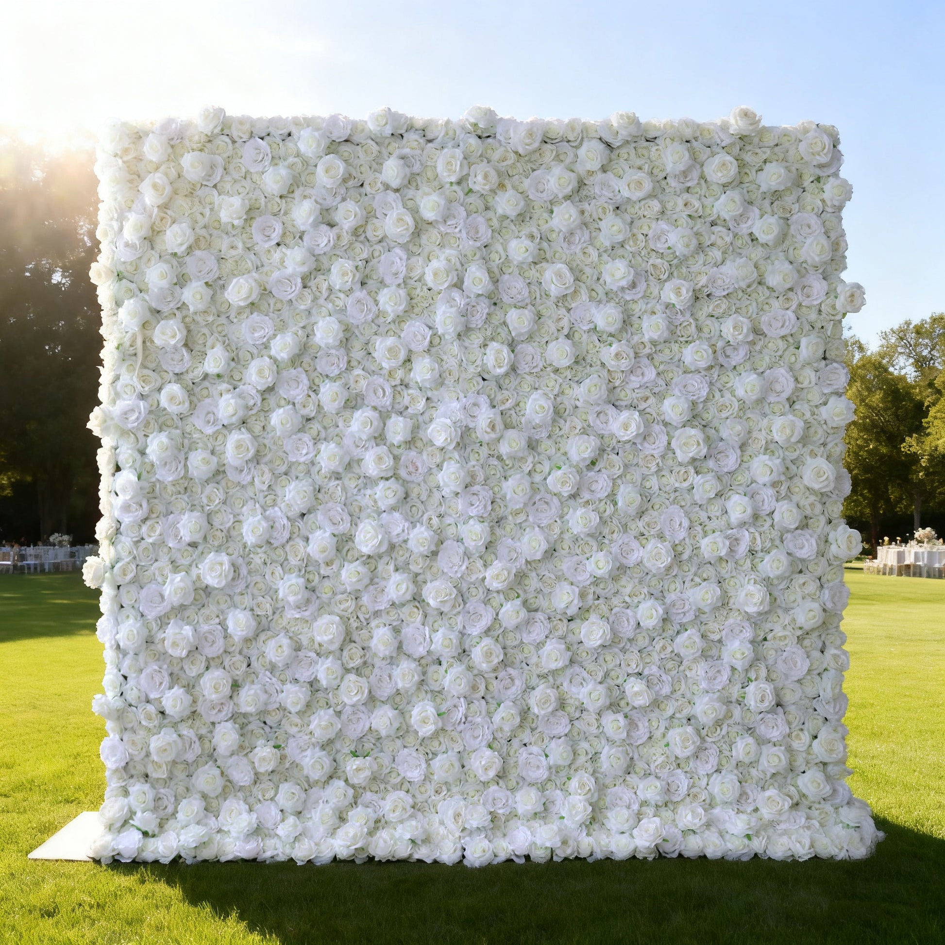 This image features a large, square floral wall outdoors on a lush green lawn. It's densely covered with pure white flowers, creating a clean, elegant, and serene display. Bathed in sunlight, with trees and event - like setups in the background, the scene exudes a sense of purity, grace, and natural beauty, perfect for decorative purposes in special events or as a captivating backdrop.