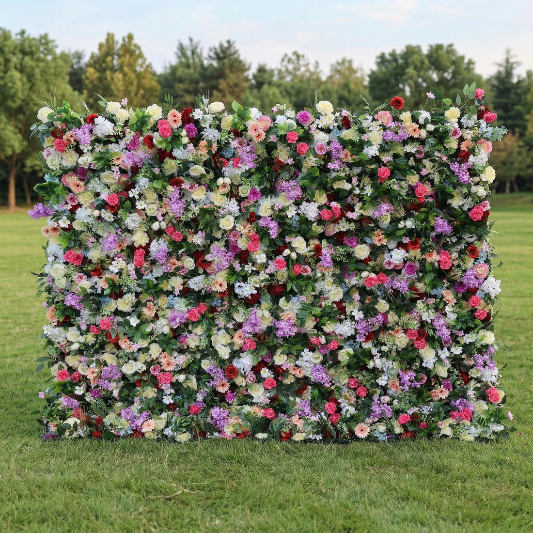 A sprawling outdoor floral wall by Valar Flowers sits on lush green grass, backed by tall trees. Brimming with vivid red roses, soft pink peonies, purple stock, creamy white blooms, and peach accents, it’s densely packed with lush green foliage. Ideal for garden weddings, outdoor receptions, engagement shoots, and festive event photo backdrops.