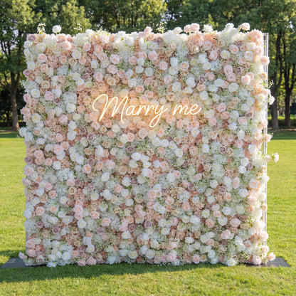 This outdoor backdrop (set on lush green grass, tree-framed) features a soft, dense wall of blush, ivory, and pale pink blooms—roses, peonies, and baby’s breath—creating an airy, romantic texture. A glowing "Marry Me" neon sign (cursive, light-lit) anchors the center, its gentle glow contrasting with the muted, elegant palette. The delicate display exudes warmth, perfect for outdoor proposals, garden weddings, or intimate romantic events.