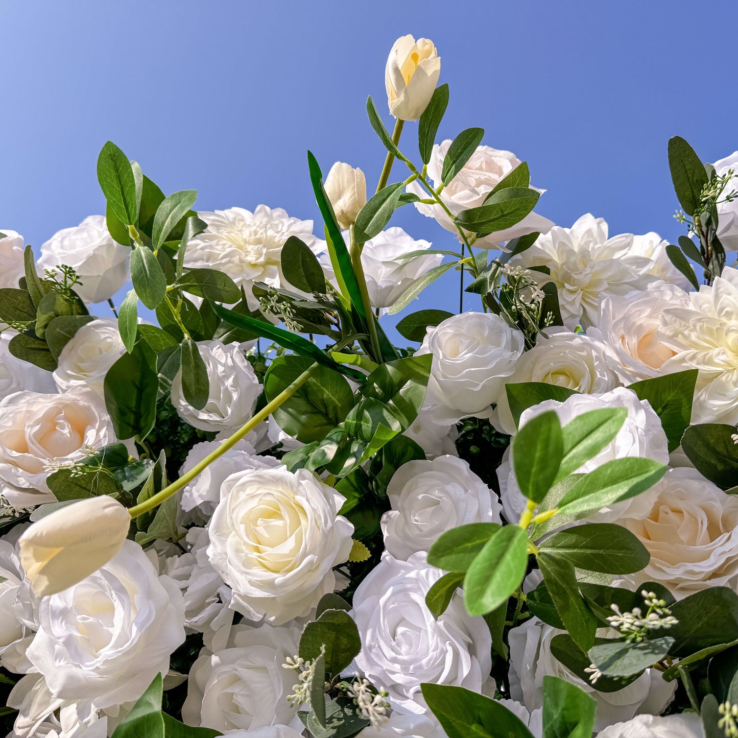 This photo showcases a lush arrangement of white roses and tulips, complemented by green foliage, set against a clear blue sky. The flowers create an elegant and serene display. Ideal for events like a “White - themed Garden Soirée” or a refined wedding, it exudes purity and sophistication, perfect for upscale celebrations. 