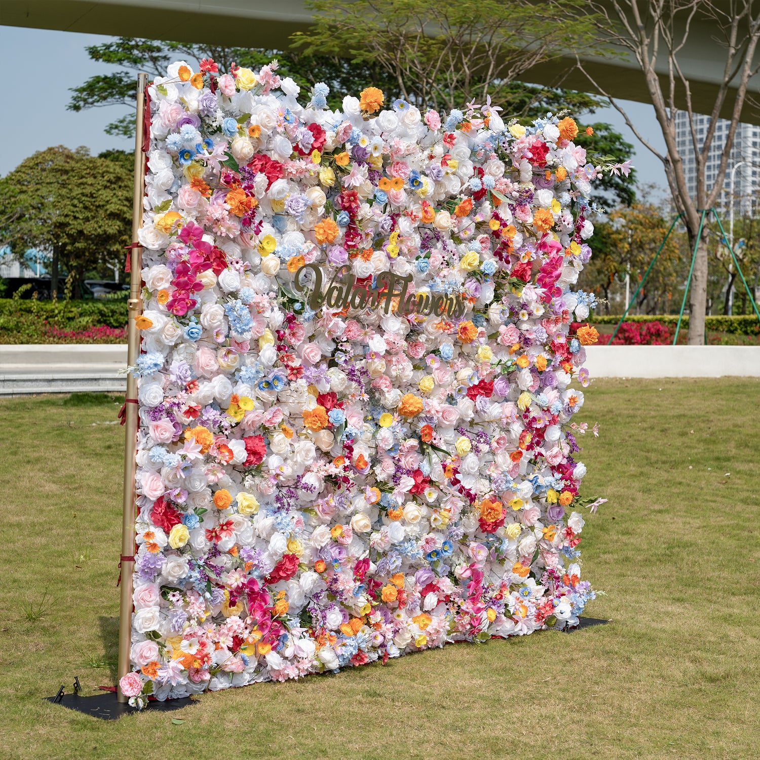 This is a breathtaking, ultra-dense artificial flower wall backdrop from Valar Flowers, set outdoors on a lush green lawn with modern urban architecture in the distance. It’s a vibrant explosion of lifelike blooms in a rainbow palette—creamy white roses, soft pink peonies, sunny yellow poppies, vivid orange carnations, sky-blue delphiniums, magenta hydrangeas, and lavender orchids. The lush, layered arrangement creates a showstopping, romantic focal point perfect for outdoor weddings, engagement parties, ph