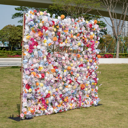 This is a breathtaking, ultra-dense artificial flower wall backdrop from Valar Flowers, set outdoors on a lush green lawn with modern urban architecture in the distance. It’s a vibrant explosion of lifelike blooms in a rainbow palette—creamy white roses, soft pink peonies, sunny yellow poppies, vivid orange carnations, sky-blue delphiniums, magenta hydrangeas, and lavender orchids. The lush, layered arrangement creates a showstopping, romantic focal point perfect for outdoor weddings, engagement parties, ph