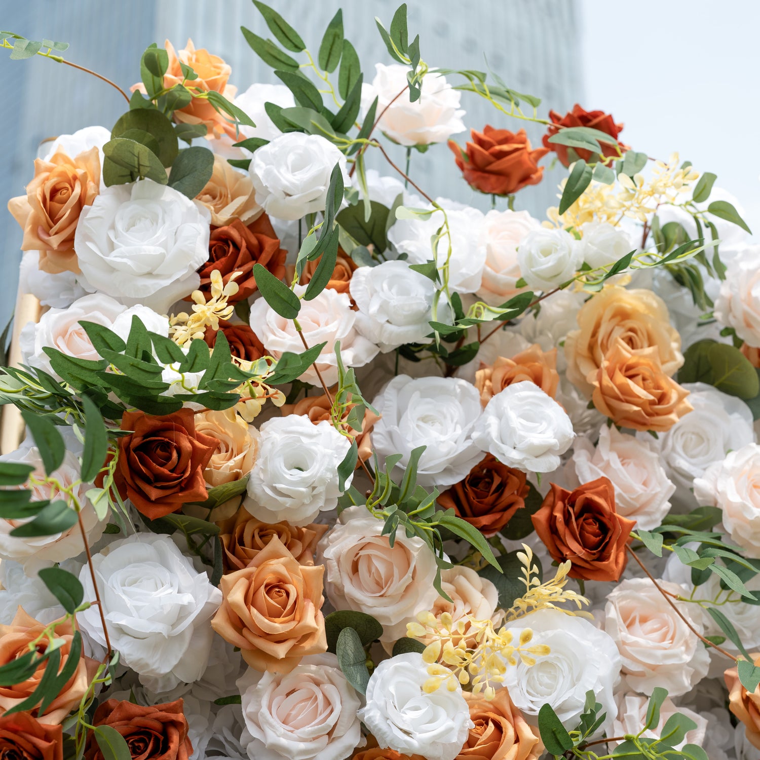 This is a lush, premium artificial flower arrangement brimming with lifelike blooms in a warm, earthy palette. It features creamy white roses, burnt orange roses, soft peach and blush roses, delicate yellow filler flowers, and realistic eucalyptus greenery. The dense, layered design creates a fresh, timeless, romantic aesthetic, perfect for wedding backdrops, photo booths, fall events, and luxury event styling.