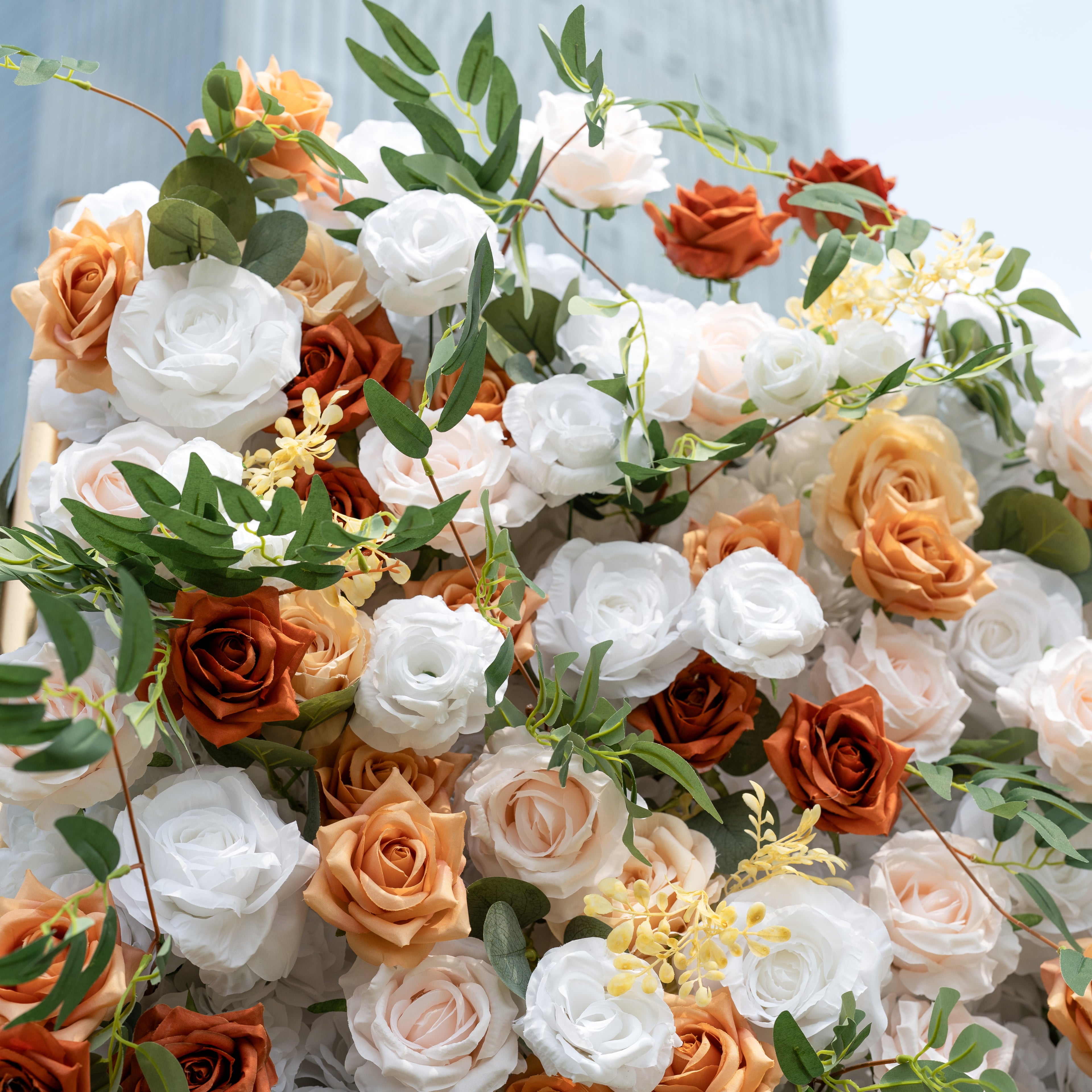 This is a lush, premium artificial flower arrangement brimming with lifelike blooms in a warm, earthy palette. It features creamy white roses, burnt orange roses, soft peach and blush roses, delicate yellow filler flowers, and realistic eucalyptus greenery. The dense, layered design creates a fresh, timeless, romantic aesthetic, perfect for wedding backdrops, photo booths, fall events, and luxury event styling.
