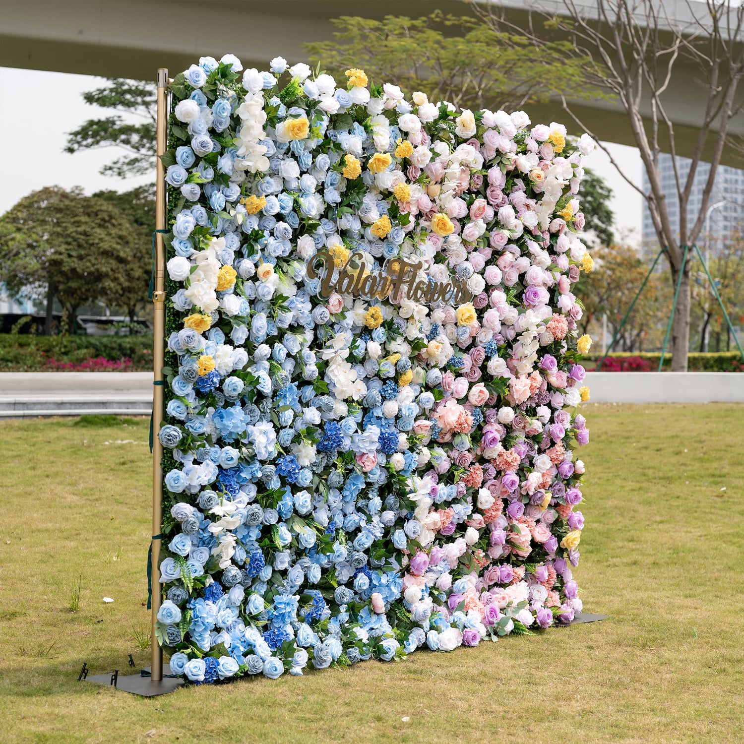 ValarFlowers’ premium gradient artificial flower wall backdrop is a lush, dense outdoor display set on a grassy lawn with an urban skyline backdrop. It features a seamless color transition from sky-blue delphiniums at the base to creamy white roses, sunny yellow poppies, blush pink peonies, and lavender roses at the top. This lifelike, reusable floral arrangement creates an elegant, romantic focal point ideal for weddings, photo booths, baby showers, and luxury event decor.