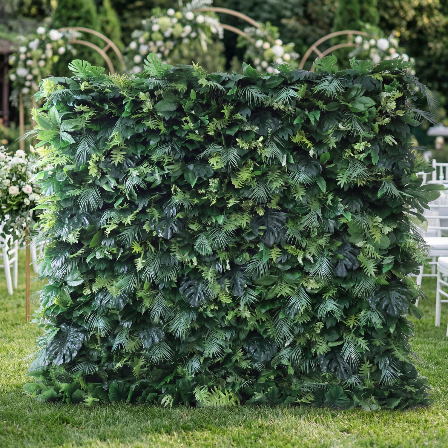 This Valar Flowers installation centers a lush, dense greenery wall—layered with varied foliage (monstera leaves, ferns, tropical fronds) in rich, deep green tones. Set on a grassy outdoor venue, a soft-focus floral arch (white blooms, gold frames) peeks behind, while white chairs flank the wall. The design feels lush and organic, blending tropical texture with elegant event styling—ideal for backdrops or decor accents, embodying Valar’s nature-inspired, cohesive aesthetic.