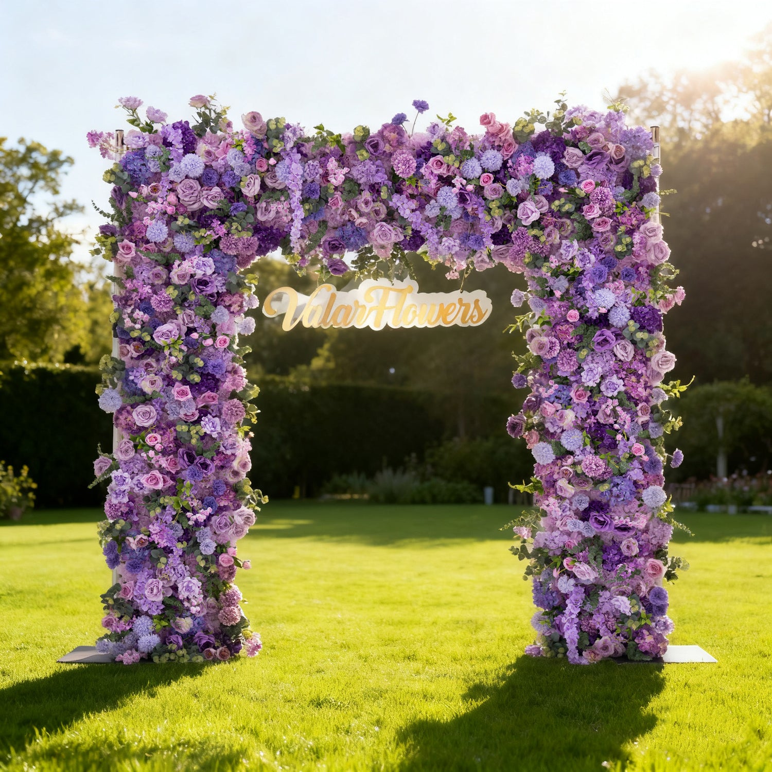 This image showcases a stunning floral arch by Valar Flowers, set outdoors on a lush green lawn. It features a dense arrangement of artificial flowers in various shades of purple, lavender, and pink, creating an elegant, nature-inspired display. The brand’s name is elegantly integrated in gold, with warm sunlight and greenery in the background, perfect for adding a luxurious touch to outdoor events like weddings.