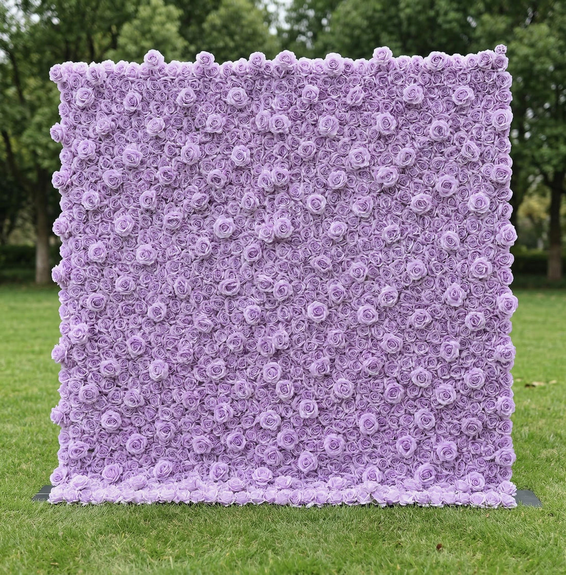 A stunning, monochromatic lavender floral wall by Valar Flowers stands on lush green grass, backed by trees. Densely packed with uniform, soft lavender roses, it creates a dreamy, textured surface. This elegant, cohesive design is perfect for romantic wedding backdrops, engagement shoots, or pastel-themed event decor, radiating a serene, sophisticated charm.