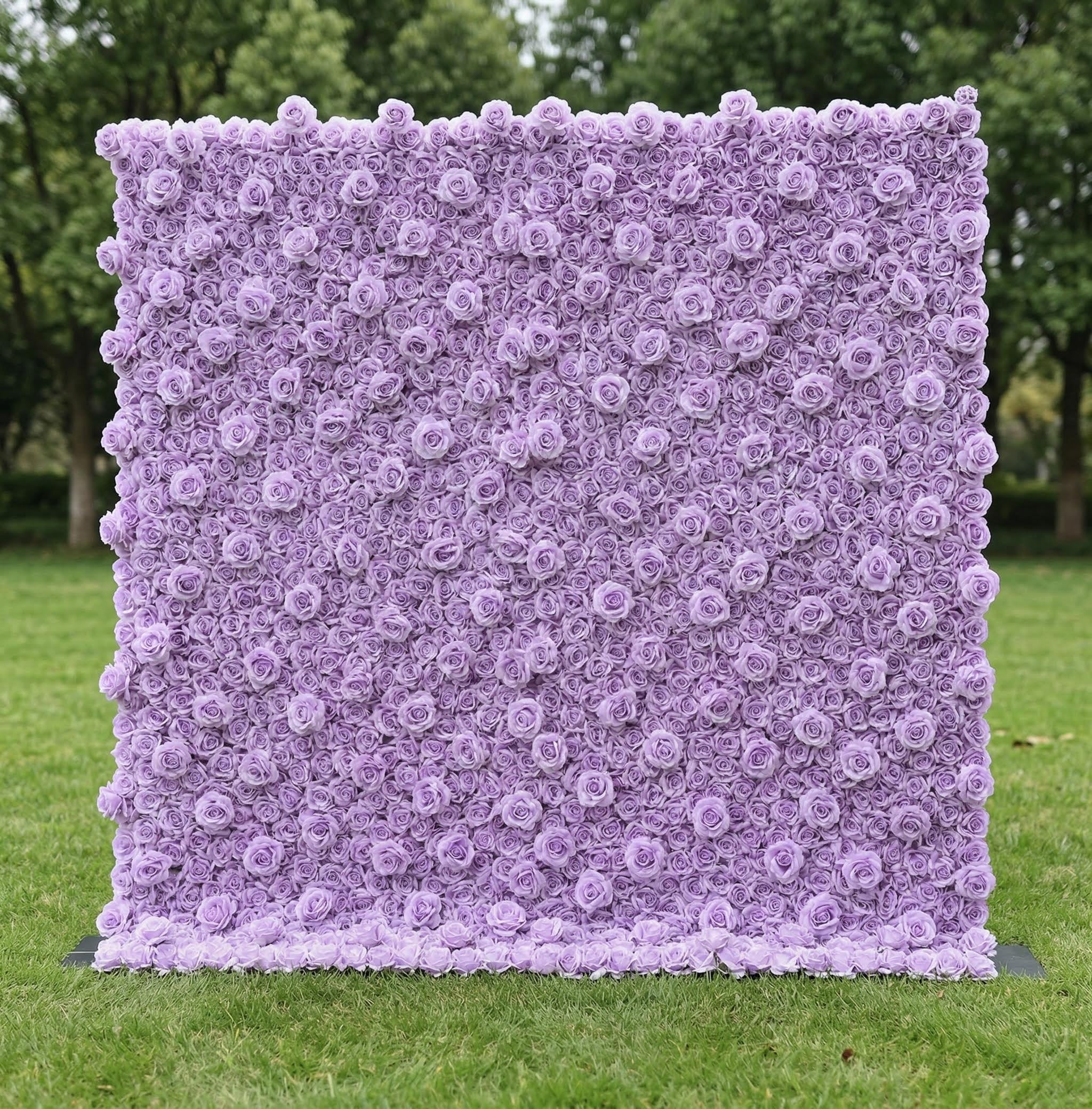 A stunning, monochromatic lavender floral wall by Valar Flowers stands on lush green grass, backed by trees. Densely packed with uniform, soft lavender roses, it creates a dreamy, textured surface. This elegant, cohesive design is perfect for romantic wedding backdrops, engagement shoots, or pastel-themed event decor, radiating a serene, sophisticated charm.
