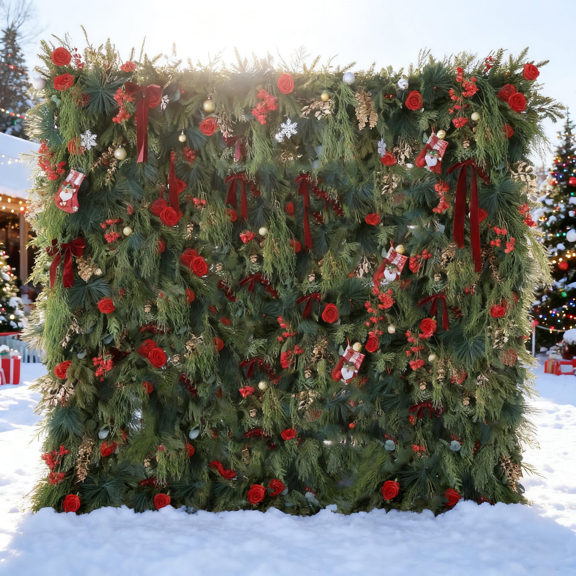 This image features a large, festive Christmas - themed floral wall. Adorned with lush green foliage, vibrant red roses, and Christmas decorations like stockings, snowflakes, and baubles, it's set in a snowy outdoor scene. In the background, lit Christmas trees enhance the joyful, wintry holiday atmosphere.