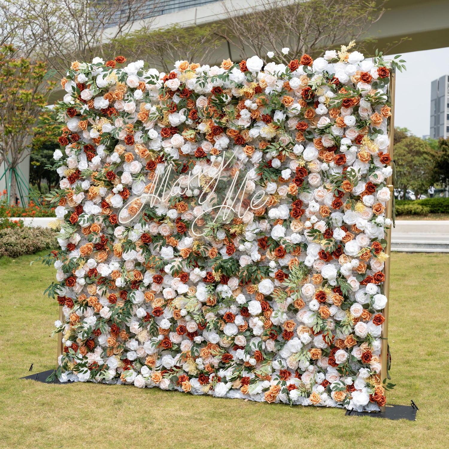 This is a stunning full-size artificial flower wall backdrop, set outdoors on a grassy lawn with an urban skyline in the background. It features a lush, dense arrangement of lifelike blooms in a warm terracotta and cream palette: burnt orange roses, creamy white peonies, soft peach roses, and vibrant green foliage, centered with an elegant "Marry Me" neon script sign. This premium, reusable floral display creates a romantic, showstopping focal point, perfect for wedding proposals, rustic weddings, fall even