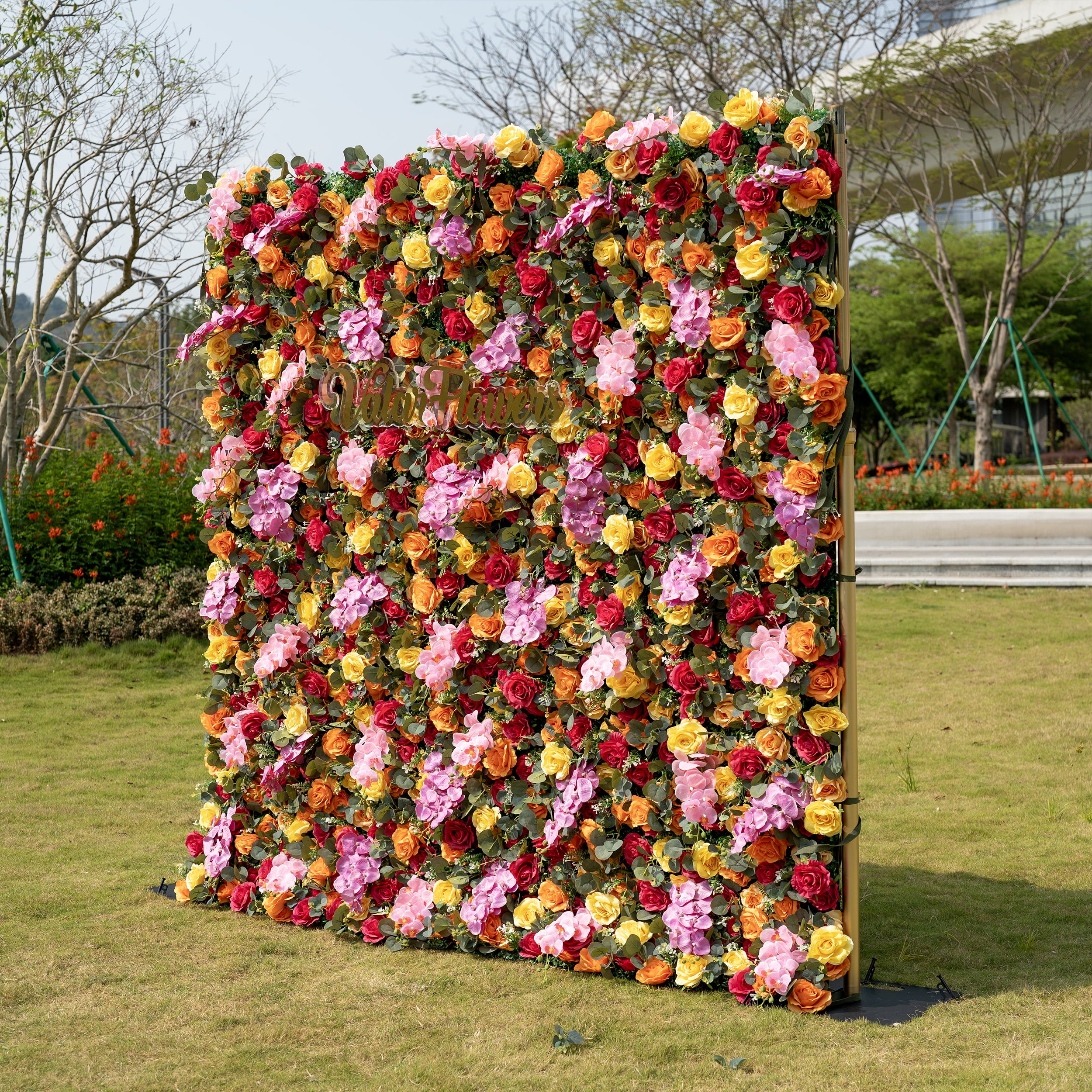 This is a full-size ValarFlowers premium multicolor artificial flower wall backdrop, set against a sunlit outdoor lawn. It features a lush, dense arrangement of lifelike burgundy, orange, yellow roses, pink hydrangeas, and eucalyptus greenery, mounted on an elegant gold adjustable stand. This high-quality, reusable floral display creates a vibrant, romantic focal point, perfect for wedding backdrops, photo booths, garden parties, and luxury outdoor event styling.