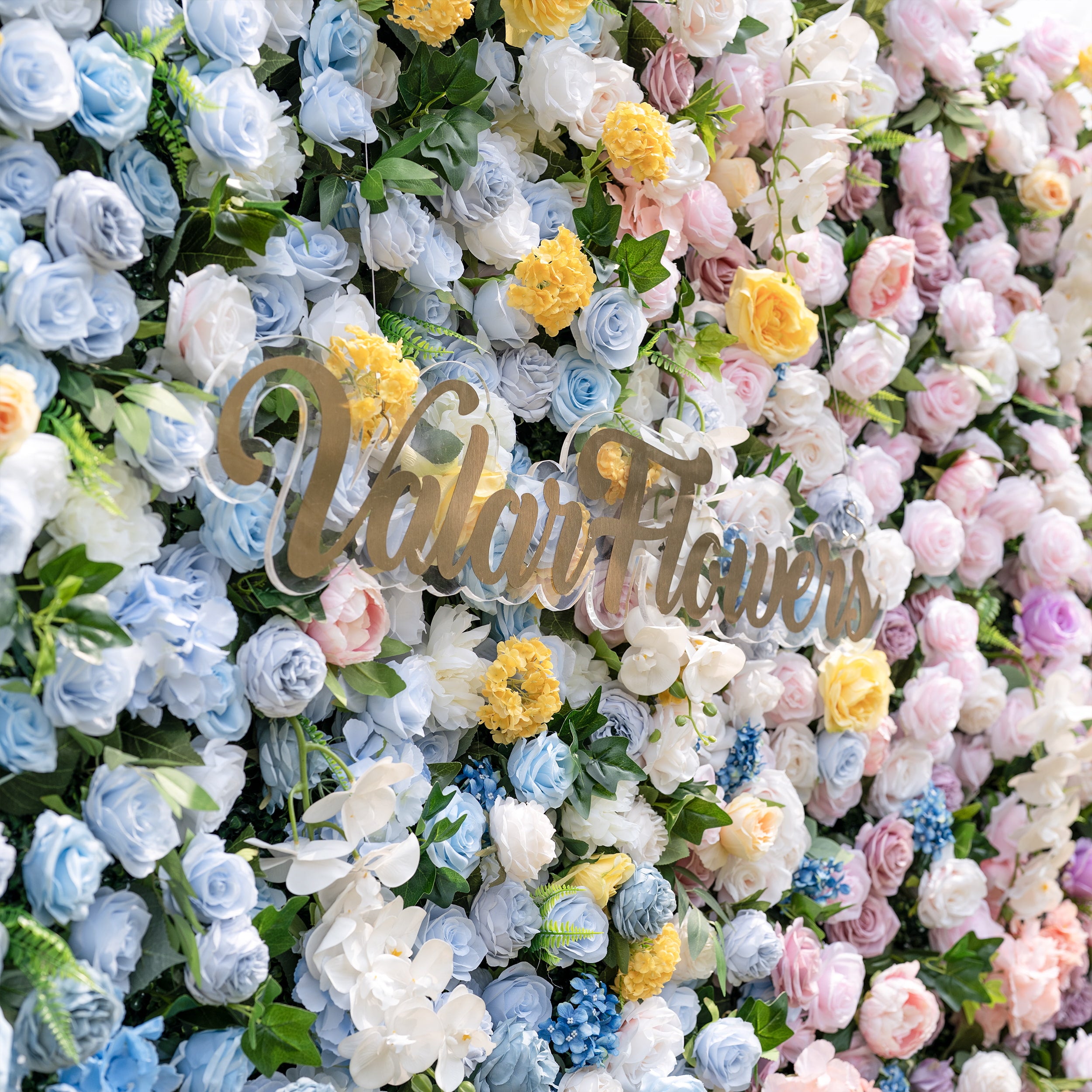 This is a lush, vibrant close-up of ValarFlowers’ premium artificial flower wall, brimming with lifelike blooms in a dreamy pastel gradient. It features sky-blue roses, creamy white peonies, sunny yellow carnations, blush pink roses, and delicate filler flowers, with the brand’s gold script logo at the center. The dense, realistic arrangement creates an elegant, romantic focal point perfect for weddings, photo booths, baby showers, and luxury event decor.
