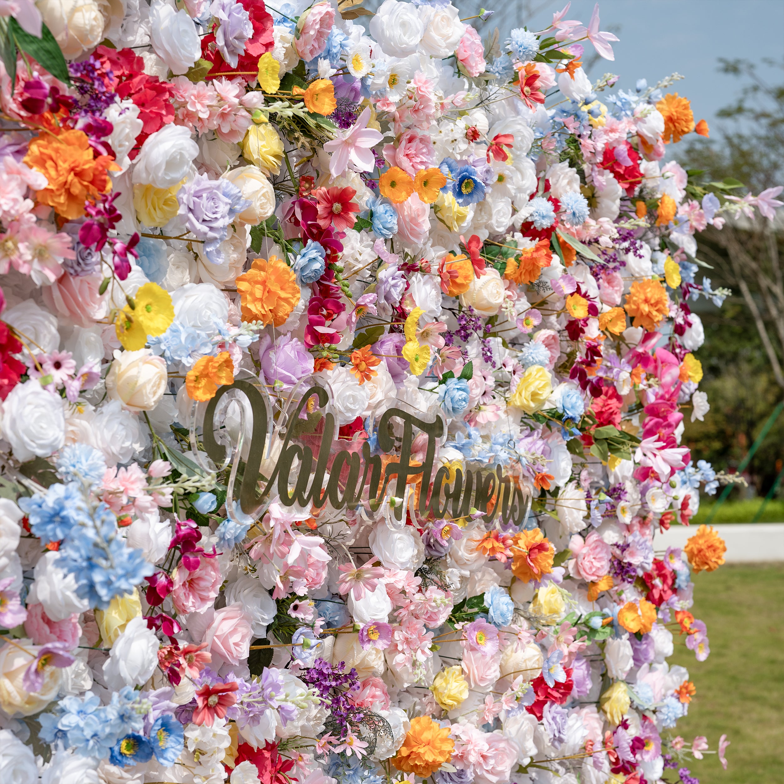 This is a lush, vibrant close-up of Valar Flowers’ premium artificial mixed-flower wall, brimming with lifelike blooms in a joyful rainbow palette. It features creamy white roses, soft pink peonies, sunny yellow poppies, vivid orange carnations, sky-blue delphiniums, deep magenta hydrangeas, lavender roses, and delicate filler flowers, set against a bright outdoor backdrop. The layered, realistic petals create a dreamy, romantic, showstopping focal point perfect for weddings, photo booths, and luxury event 