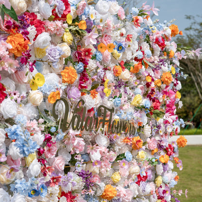 This is a lush, vibrant close-up of Valar Flowers’ premium artificial mixed-flower wall, brimming with lifelike blooms in a joyful rainbow palette. It features creamy white roses, soft pink peonies, sunny yellow poppies, vivid orange carnations, sky-blue delphiniums, deep magenta hydrangeas, lavender roses, and delicate filler flowers, set against a bright outdoor backdrop. The layered, realistic petals create a dreamy, romantic, showstopping focal point perfect for weddings, photo booths, and luxury event 