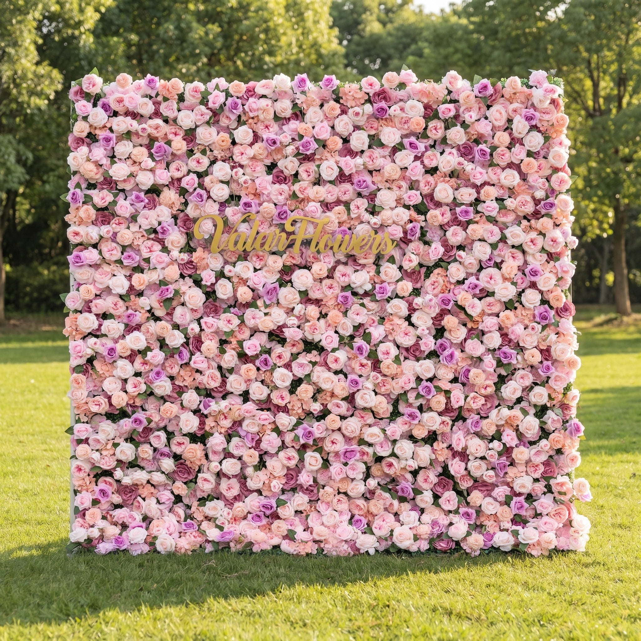 Valar Flowers Sunset Romance flower wall front view with blush pink dusty rose peach and ivory silk roses peonies and hydrangeas accented by gold script sign on green lawn under bistro lights for romantic wedding backdrops and luxury event decor