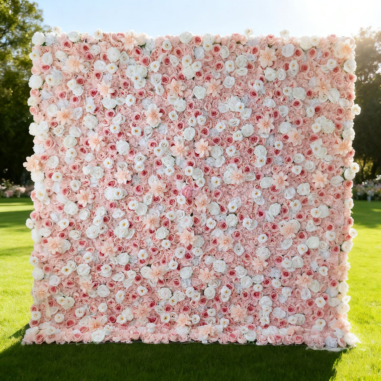 This image features a large, square floral wall outdoors on a lush green lawn. It's densely covered with an array of flowers in soft pink, white, and hints of red. The vibrant blooms create a rich, colorful, and romantic display. Bathed in sunlight, with trees and a clear sky in the background, the scene exudes a sense of beauty, elegance, and natural charm, perfect for decorative purposes in events or as a picturesque backdrop.