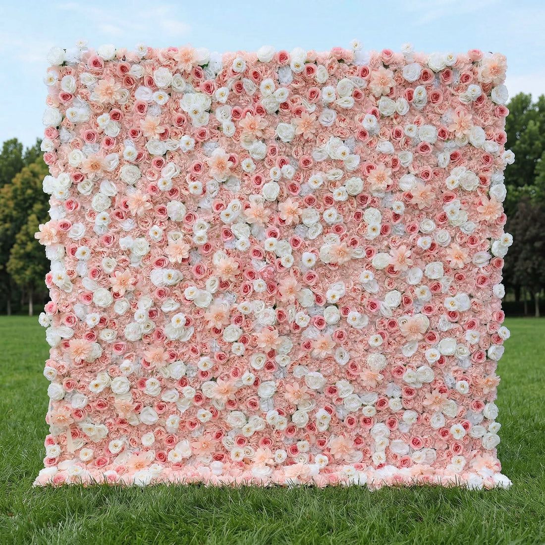 A dreamy, romantic floral wall by Valar Flowers stands on lush green grass, backed by trees. Densely packed with a soft gradient of blush pink, creamy white, and peach blooms—including roses, peonies, and delicate daisies—it creates a seamless, textured surface. This elegant, pastel design is perfect for garden weddings, engagement shoots, or feminine-themed event photo backdrops, radiating warmth and sophistication.
