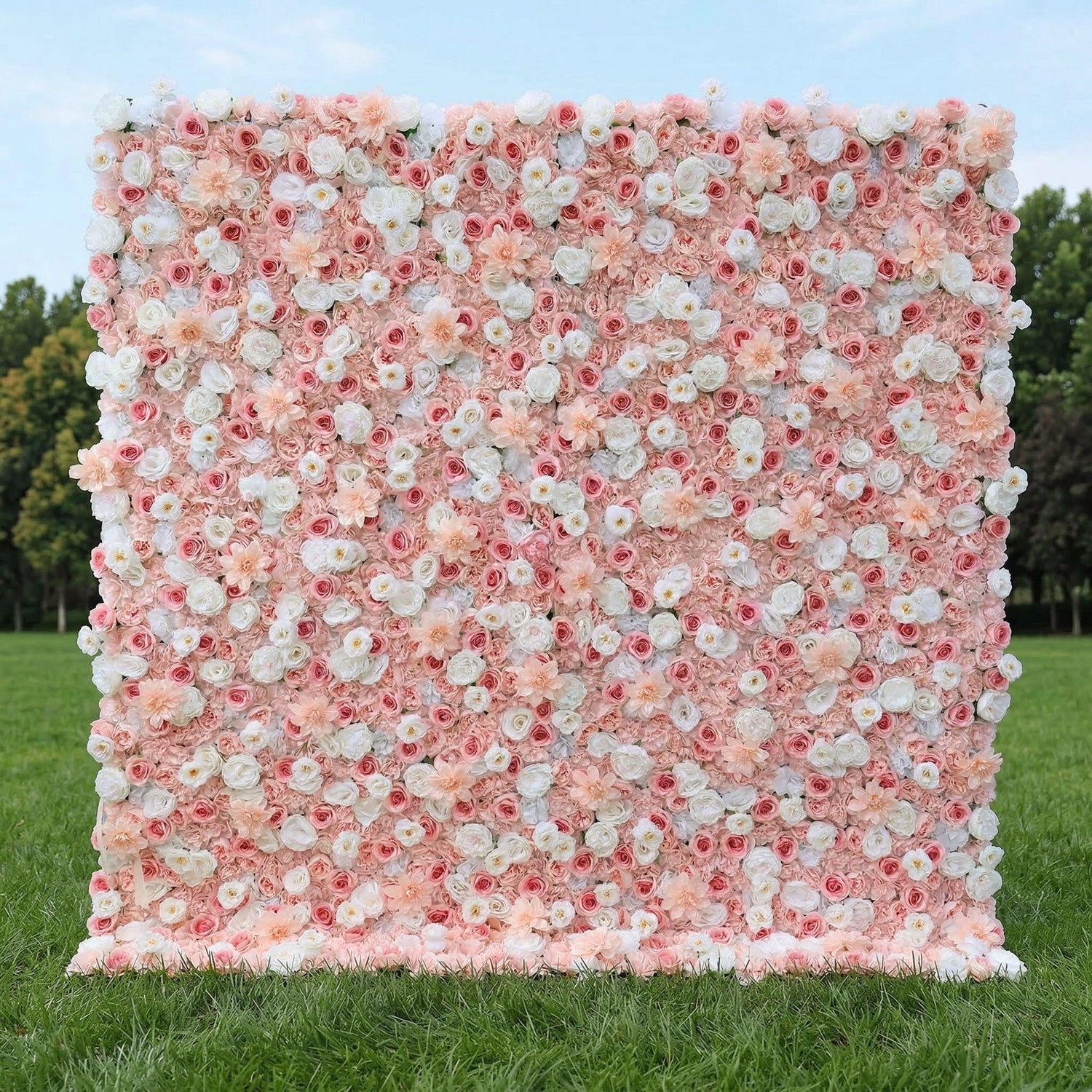A dreamy, romantic floral wall by Valar Flowers stands on lush green grass, backed by trees. Densely packed with a soft gradient of blush pink, creamy white, and peach blooms—including roses, peonies, and delicate daisies—it creates a seamless, textured surface. This elegant, pastel design is perfect for garden weddings, engagement shoots, or feminine-themed event photo backdrops, radiating warmth and sophistication.