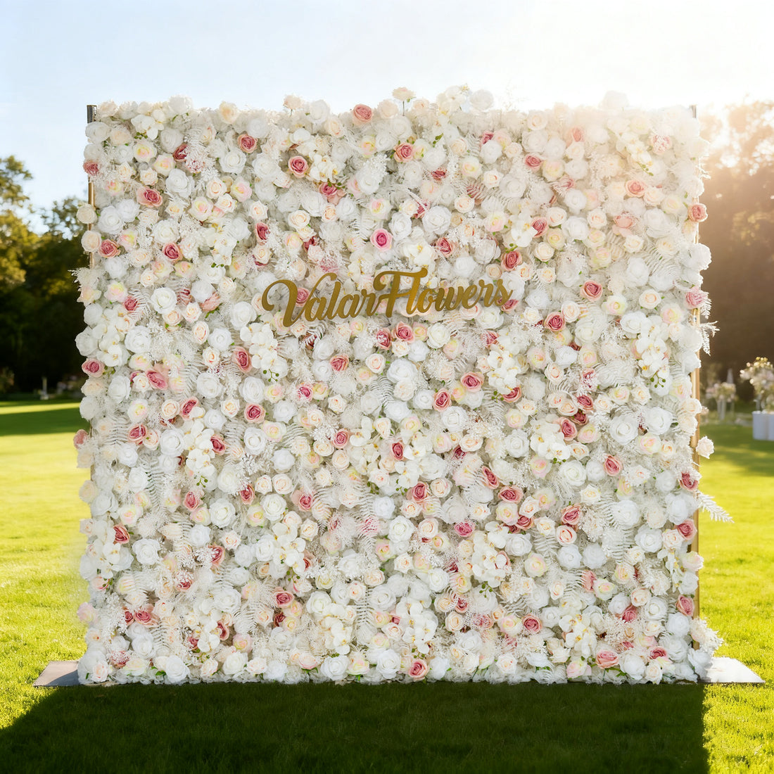 This image showcases a large floral wall densely covered with white flowers, accented with hints of pink. &quot;Valar Flowers&quot; is displayed in golden text. Set outdoors on a lush green lawn under a bright sky with sunlight casting a warm glow, it creates an elegant and serene atmosphere, perfect for events or decorative displays, exuding a sense of natural beauty and refinement.