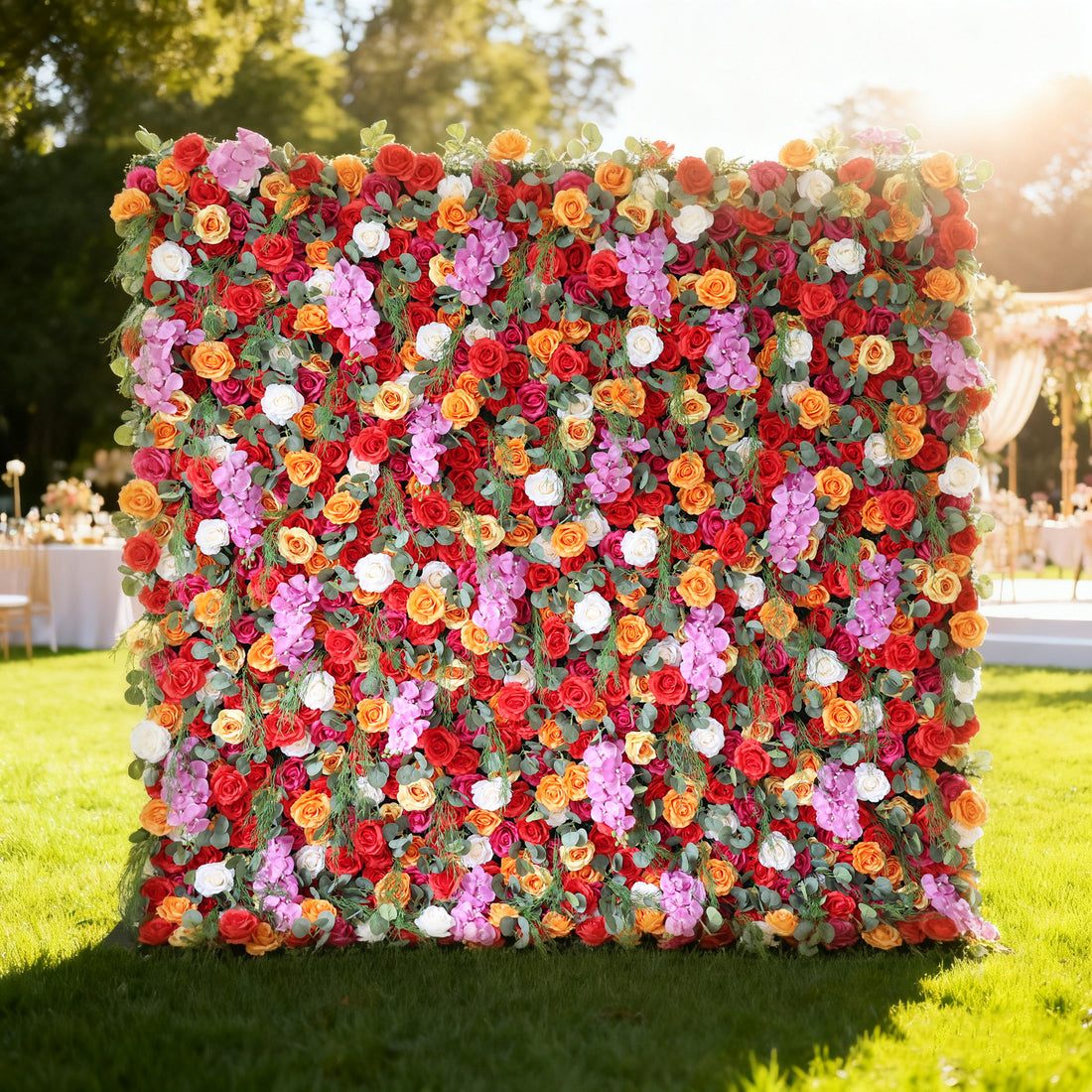 This image showcases a vibrant floral wall by Valar Flowers, set outdoors on a lush green lawn. It features a dense arrangement of artificial flowers in a rich palette of red, orange, pink, and white, creating a lively and eye-catching display. The backdrop is perfect for events, with the bold hues and lush texture adding a touch of energy and elegance, complemented by the sunny outdoor setting and event furniture in the background.