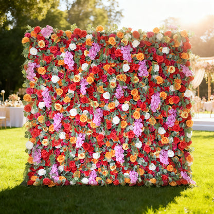 This image showcases a vibrant floral wall by Valar Flowers, set outdoors on a lush green lawn. It features a dense arrangement of artificial flowers in a rich palette of red, orange, pink, and white, creating a lively and eye-catching display. The backdrop is perfect for events, with the bold hues and lush texture adding a touch of energy and elegance, complemented by the sunny outdoor setting and event furniture in the background.