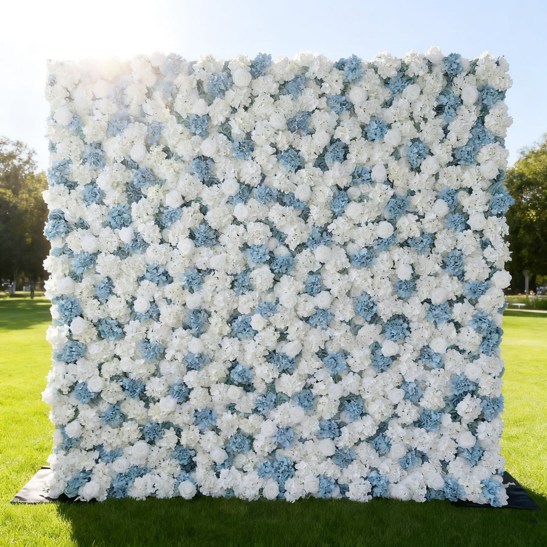 This image features a large, square floral wall composed of white and light blue flowers. Set outdoors on a lush green lawn under a bright sky, the dense arrangement of blooms creates a soft, elegant, and refreshing look. It&