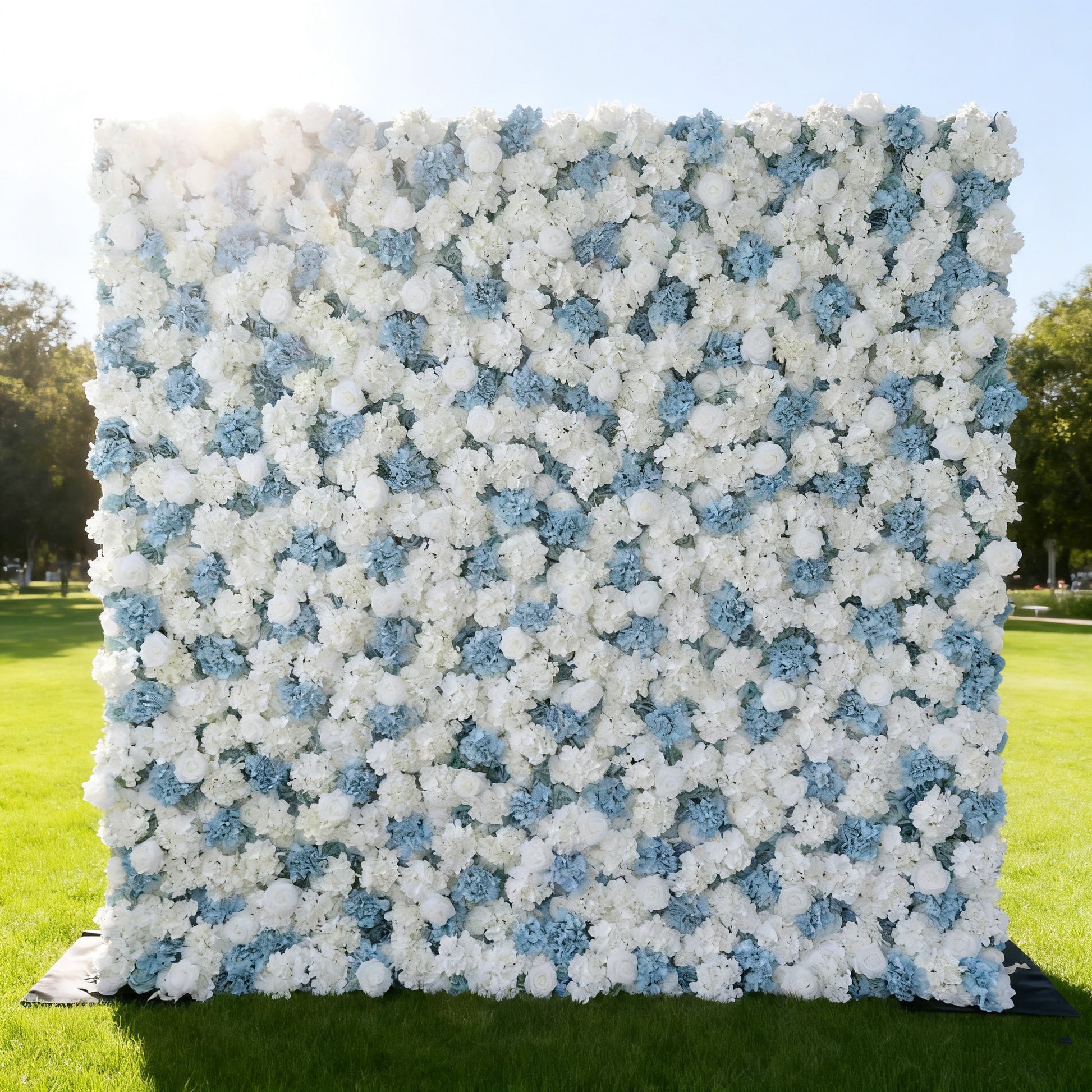 This image features a large, square floral wall composed of white and light blue flowers. Set outdoors on a lush green lawn under a bright sky, the dense arrangement of blooms creates a soft, elegant, and refreshing look. It's perfect for decorative purposes, such as events or photo backdrops, exuding a serene and charming atmosphere.