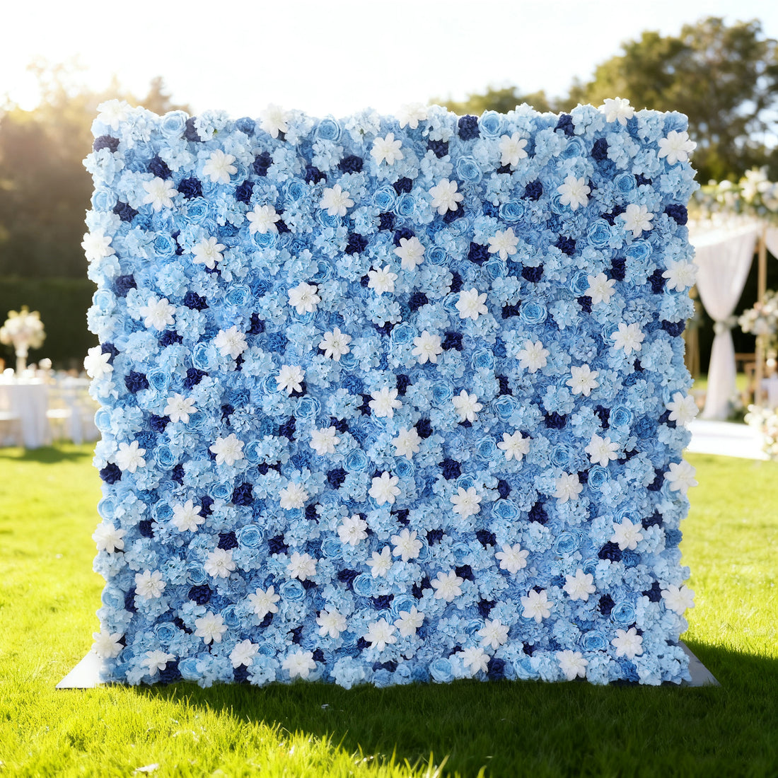 This image showcases a beautiful floral wall by Valar Flowers, set outdoors on a lush green lawn. It features a dense arrangement of artificial flowers in various shades of blue and white, creating a serene and elegant display. The backdrop is perfect for events, with the soft hues and lush texture adding a touch of sophistication, complemented by the sunny outdoor setting and event furniture in the background.